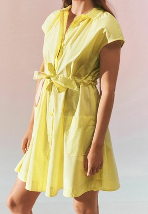 Femme portant une robe jaune à manches courtes avec des boutons sur le devant, une ceinture à la taille et de grandes poches, debout devant un fond rose clair et gris.