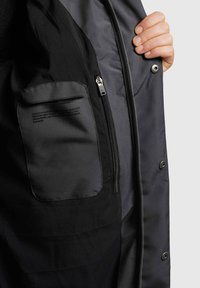 Mustan jope sisemus, millel on tõmblukuga tasku ja kangast silt koos tekstiga. Materjal on sile ja vastupidav, matt viimistlus.