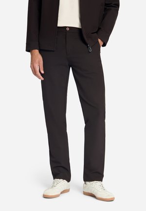 Mann trägt schwarze Hose, weiße Sneakers, weißes Hemd und schwarze Jacke, steht mit einer Hand in der Tasche vor weißem Hintergrund.