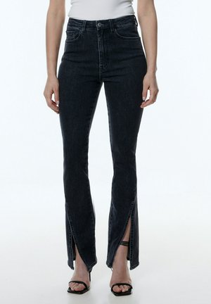 Jean en denim noir taille haute avec ourlets fendus ; comporte cinq poches et une fermeture à bouton ; texture lisse et design ajusté.