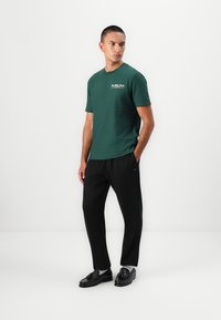 Grøn bomulds T-shirt med rund hals og korte ærmer, kombineret med sorte bukser og sorte sko. Har logo på brystet.