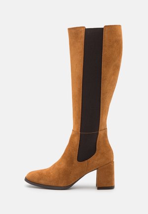 Bottes au genou en daim beige avec un panneau en élastique noir. Le bout carré et le talon bloc offrent de la stabilité. La texture lisse contraste avec l'élastique.
