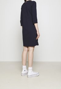 Marinblå klänning med trekvartslånga ärmar, slät textur, knälång, kombinerad med vita högskåp-sneakers och manschetter. Brun etikett vid halsringningen.