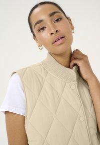 Gilet beige matelassé avec un col côtelé et des boutons-pression, fabriqué à partir d'un tissu léger. Porté par-dessus un t-shirt blanc.