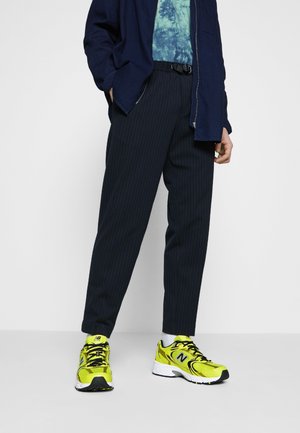 Homme portant un pantalon bleu marine à fines rayures, une veste bleu foncé, une chemise tie-dye bleue et des baskets New Balance jaune vif, debout devant un fond blanc.