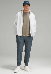 Szara bluza z kapturem zapinana na zamek, nałożona na oliwkową koszulkę, w połączeniu z ciemnoniebieskimi spodniami i białymi trampkami. Dodatki to granatowa czapka.