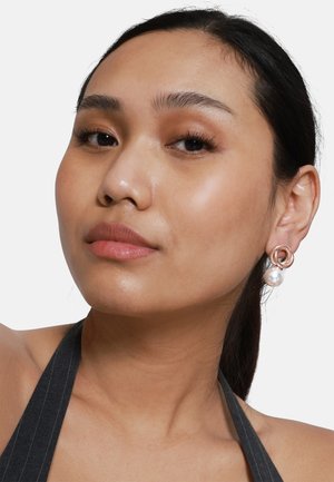Ohrringe mit einem kreisförmigen roségoldenen Design und einer Perle, die herabhängt. Das Modell hat langes dunkles Haar und einen glatten Teint und trägt ein dunkles Neckholder-Oberteil.