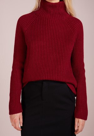 Femme portant un pull à col roulé rouge côtelé à manches longues, rentré dans une jupe noire taille haute sur un fond neutre.