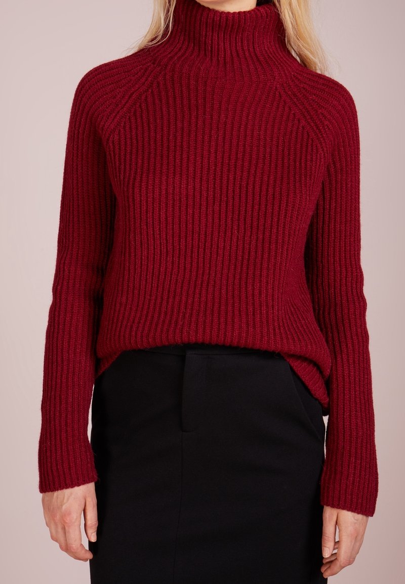 Femme portant un pull à col roulé rouge côtelé à manches longues, rentré dans une jupe noire taille haute sur un fond neutre.