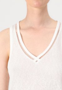 Biely top s výstrihom do V a výrezmi pozdĺž výstrihu, vyrobený z ľahkého materiálu. Bezrukávový dizajn s mäkkou textúrou.