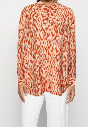 Woman wearing long-sleeve button-up shirt with red and beige abstract pattern, paired with white pants, standing against plain background.