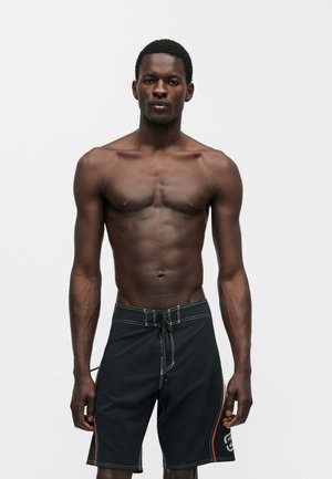 Mężczyzna o ciemnej karnacji bez koszulki, ubrany w czarne kąpielówki z białymi przeszyciami i pomarańczowymi bocznymi paskami, stojący na jednolitym jasnym tle.