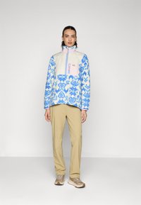 Sudadera de forro polar con patrón floral en azul y blanco, frente acolchado beige, cremallera rosa, combinada con pantalones beige y zapatillas de deporte marrón claro.