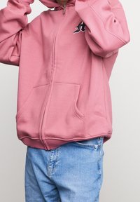 Sudadera con cremallera rosa hecha de un material suave, con un bolsillo tipo canguro y un logo en contraste en el pecho, combinada con jeans de color azul claro.