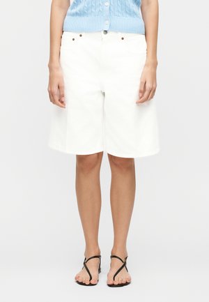 Άτομο που φοράει λευκά σορτς έως το γόνατο, ανοιχτό μπλε πλεκτό τοπ και μαύρα φλατ σανδάλια με λεπτά λουράκια.