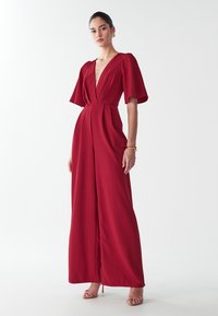 Vrouw in een dieprode wijde jumpsuit met fladdermouwen, V-hals, gouden armband, oorbellen en naakte, gespsandalen, staande op een witte achtergrond.