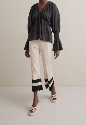 Blusa oscura de manga larga con puños fruncidos combinada con pantalones crema acanalados con franjas negras plisadas en el dobladillo y sandalias beige de plataforma.