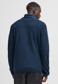 Pullover in pile blu navy con colletto alto e maniche lunghe, caratterizzato da una texture liscia e un design semplice, indossato con jeans scuri.