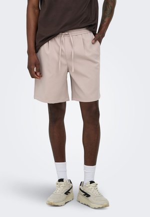 Homme portant un short beige à cordon, des chaussettes blanches montantes et des baskets beige avec des accents noirs, debout avec une main dans la poche.