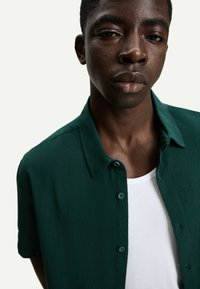 Green textured short-sleeve shirt with a buttoned collar, layered over a white tank top. Features a subtle crinkled fabric.