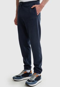 Muž oblečený v tmavě modrých teplákových kalhotách s elastickými manžetami a tmavě modrých a oranžových teniskách, stojící s jednou rukou v kapse.