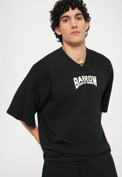 T-shirt noir à manches courtes avec un col en V, comportant le texte blanc "BARROW HALL OF FAME" à l'avant et un accent jaune près du cou.