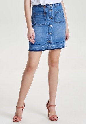 Femme portant une jupe en denim bleue boutonnée à l'avant avec des poches avant et des sandales à talons hauts roses, debout devant un fond uni.