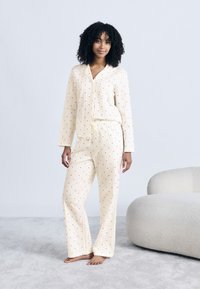 Γυναίκα με σγουρά μαλλιά που στέκεται ξυπόλητη σε χαλί, φορώντας κρεμ πιτζάμες με μικρό κόκκινο σχέδιο καρδιών, δίπλα σε λευκή επενδεδυμένη καρέκλα.