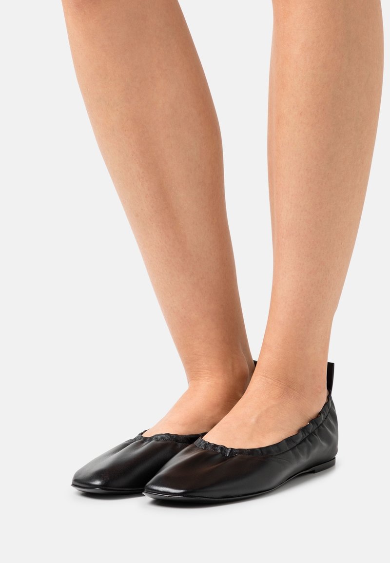 Ballerines en cuir noir portées pieds nus, vues de profil sur un fond clair uni.