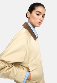 Kobieta z wygładzeniem włosów nosi beżową kurtkę z brązowym kołnierzem z korduory i jasnoniebieską koszulę, ręka w kieszeni kurtki, neutralne tło.