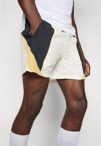 Herre sportshorts: hvide med sorte og gule detaljer, med mesh-paneler og en lomme, skåret over knæet. Letvægtsstof.