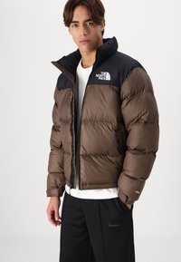 Jachetă puffer maro cu umeri din nylon negru, guler înalt, închidere cu fermoar și un logo pe pieptul stâng. Prezintă un model matlasat și izolație de 700.