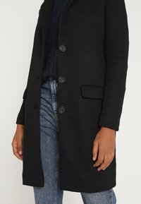 Cappotto nero lavorato a maglia con un design strutturato, dotato di chiusura a bottoni, due tasche frontali e maniche lunghe. Il tessuto testurizzato aggiunge profondità.