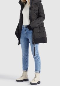 Mujer vestida con una chaqueta de plumas negra, suéter blanco de canalé, jeans azules con puños, guantes negros y botas de cordones color crema, de pie sobre un fondo blanco.