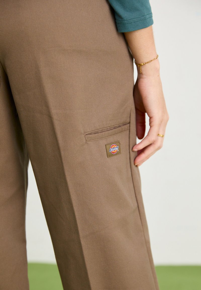 Braune weit geschnittene Hose aus strapazierfähigem Stoff, ausgestattet mit einer seitlichen Tasche und einem Logo-Patch. Die Textur wirkt glatt und hat minimale Falten.