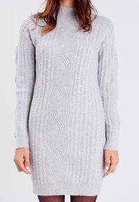 Robe en maille gris clair présentant une texture côtelée, des manches longues et un col montant. Le modèle inclut un motif ondulé sur le devant.