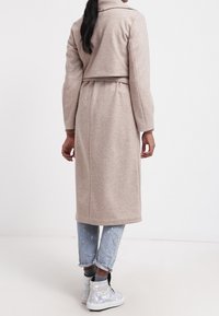 Mujer con un abrigo largo color beige sobre unos jeans azul claro remangados y zapatillas altas iridiscentes, de pie contra un fondo blanco.