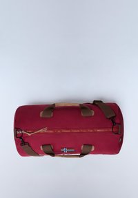 Borsa da viaggio in tessuto rosso con manici e tracolla marroni. Apertura con zip, con una toppa a forma di bandiera sul lato. Forma robusta e cilindrica.