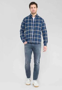 Camisa de franela en cuadros azules y grises, con cierre de botones y cuello. Llevada con jeans grises y zapatillas blancas. La textura parece suave.