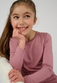 Maglia a maniche lunghe di colore rosa in un tessuto morbido; presenta un collo rotondo. Bambino con la testa appoggiata sulla mano, sorridente, con capelli lisci, seduto su una sedia bianca.