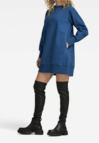 Blå oversized sweatshirtklänning med långa ärmar och sidofickor, kombinerad med knähöga svarta faux läderstövlar med kraftiga sulor.