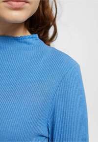 Blå ribbad långärmad topp med hög hals. Tyget är slätt med en subtil textur, och kanterna har en vågig design.