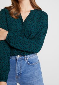 Blusa verde azulado con estampado de leopardo negro, mangas largas con puños fruncidos y escote en V, combinada con jeans de cintura alta en color azul claro.