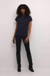 T-shirt en coton bleu marine avec des manches courtes retroussées, portée avec un jean noir ajusté et des bottes en cuir noir jusqu'aux genoux sur un fond uni.