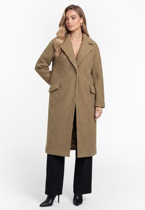 Donna che indossa un lungo cappotto marrone testurizzato con ampi rever, pantaloni neri e stivali neri a punta con tacco alto, in posa su sfondo bianco.