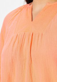 Pfirsichfarbene Bluse aus strukturiertem Stoff. Mit einem V-Ausschnitt und gerafften Details unterhalb des Ausschnitts. Weiches, leichtes Material.