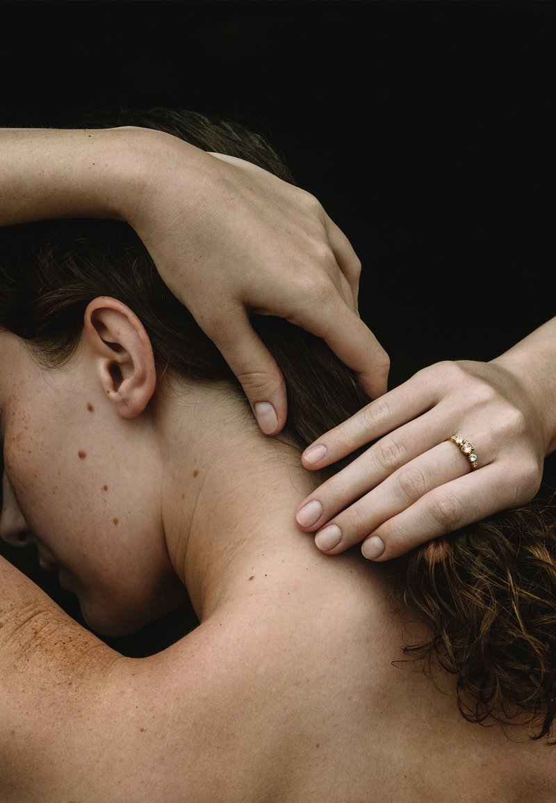 Person med bar ryg og krøllet hår, hænderne rører blidt ved nakke og hår, den ene hånd bærer en guldring med tre sten.