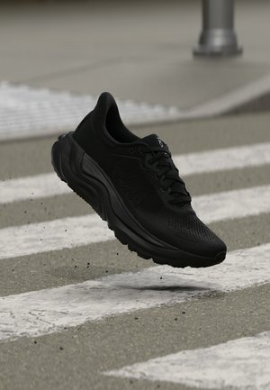Chaussure de course athlétique noire en l'air au-dessus d'un passage piéton avec une semelle texturée et une matière supérieure respirante.