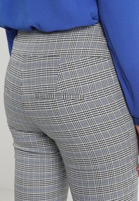 Gros plan sur une personne portant un pantalon à carreaux noir, blanc et bleu avec des poches arrière, et un haut à manches longues bleu texturé.