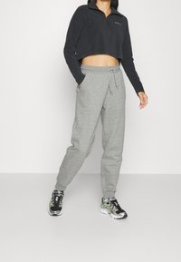 Geschnittenes schwarz meliertes Zip-up-Oberteil mit langen Ärmeln, kombiniert mit hellgrauen, zulaufenden Jogginghosen und schwarzen Sportschuhen mit grünen Akzenten.